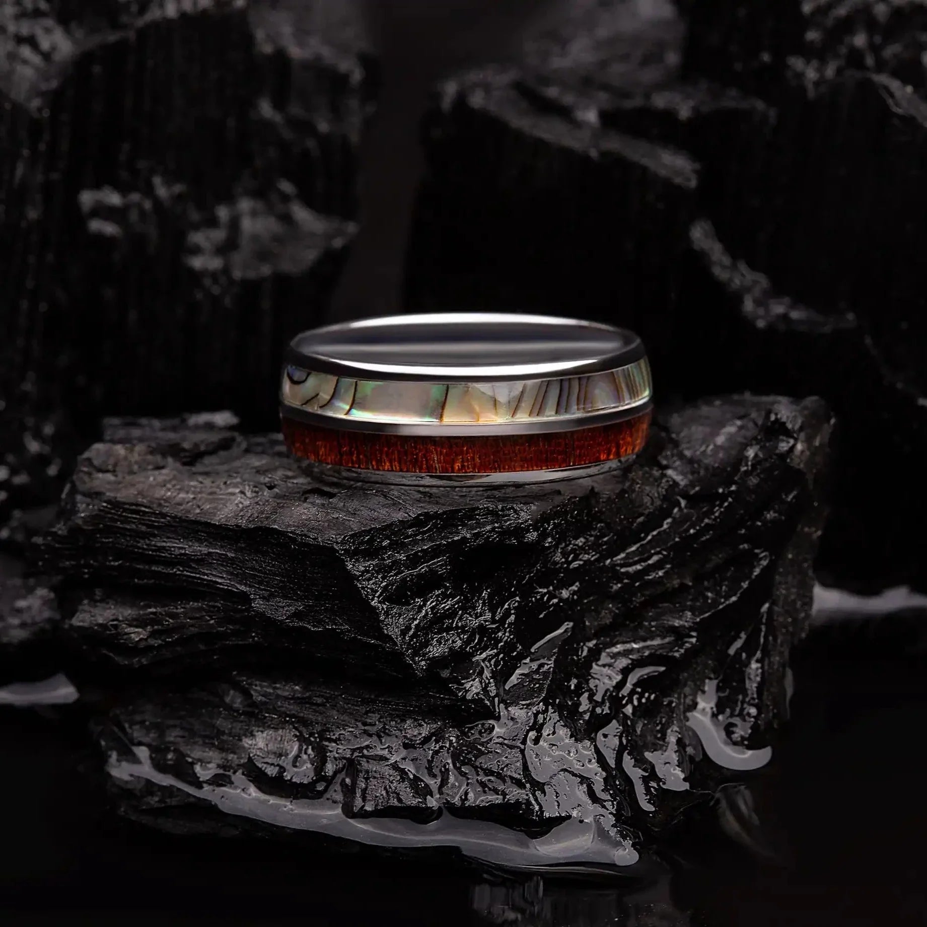 Brushed silver tungsten ring with abalone shell and koa wood. The ring is laying on a black coal